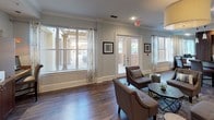 This single-floor amenity space is thoughtfully designed to serve as a welcoming communal gathering area. The open layout is anchored by rich hardwood-style flooring that flows throughout, complemented by soft light gray walls, crisp white crown molding, and recessed lighting that creates a warm, inviting ambiance.

The main lounge area features multiple seating arrangements, including tufted armchairs and a sofa positioned around a central coffee table, ideal for relaxed socializing. A wall-mounted flat-screen television sits above a warm cherry wood media cabinet, adding a residential comfort to the space. A small bistro table nearby offers an additional casual seating option.

A standout feature is the curved wet bar, finished with rich dark wood cabinetry, dark granite countertops, a mosaic tile backsplash, and elegant pendant lighting overhead — lending a sophisticated, upscale feel. Bar seating is provided for convenient use.

Natural light floods the space through expansive windows dressed with sheer curtains, while French doors open directly to an outdoor patio area, seamlessly connecting the interior to the exterior. A dedicated workstation area with counter seating rounds out the functional offerings of this well-appointed space.