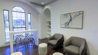 This single-floor space showcases a thoughtfully designed interior that blends classic architectural elements with contemporary styling. The open layout is anchored by striking arched details, including a graceful arched alcove housing built-in shelving — an elegant focal point that adds depth and character to the bright white walls. A large arched window along one side floods the space with natural light, offering views of lush greenery outside and enhancing the airy, welcoming atmosphere.

The seating area is furnished with sleek, modern grey armchairs complemented by a plush round ottoman, creating an inviting conversational arrangement. A large abstract artwork in warm, neutral tones adorns the wall, adding a refined artistic touch. Recessed lighting throughout provides consistent illumination, while dark hardwood flooring grounds the otherwise light and neutral palette.

A bar-height counter with industrial-style metal stools defines a secondary gathering area, offering a casual alternative for seating. The interplay of white walls, natural wood tones, metallic accents, and soft grey upholstery gives the space a polished yet approachable feel throughout.