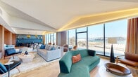 Located at 39 Bruton Street in San Francisco, CA, this single-floor space offers a generously proportioned open-plan layout with striking views of the San Francisco Bay and the iconic Bay Bridge through expansive floor-to-ceiling windows. Natural light floods the interior throughout the day, highlighting the wide-plank hardwood flooring and refined contemporary design that defines every corner of the space.

The living area is anchored by a bold teal velvet sofa and complemented by warm amber accent chairs, with a sleek modern fireplace adding an intimate focal point. Architectural cove lighting runs along the ceiling perimeter, casting a warm ambient glow that enhances the sophisticated atmosphere. A second seating area features rich wood paneling, built-in cabinetry, and a wall-mounted display screen, offering a more enclosed and cozy retreat.

The dining area is a design statement in its own right, featuring a long dark wood table surrounded by wooden chairs and illuminated by a trio of dramatic amber globe pendants. A fluted sage-green sideboard runs the length of one wall, adding texture and elegance. Floor-to-ceiling windows frame sweeping views of the bay and surrounding cityscape, seamlessly blending the interior with the stunning outdoor panorama. The overall ambiance is one of curated luxury and calm sophistication.