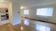 This single-floor space offers a clean and functional layout defined by its timeless architectural simplicity. The main living area is generously proportioned, featuring polished hardwood floors that reflect natural light beautifully throughout the room. Crisp white walls provide a neutral backdrop, creating a bright and airy atmosphere that feels open and welcoming.

Multiple windows fitted with horizontal blinds allow abundant natural light to pour into the space, enhancing the sense of openness. The windows are well-positioned to provide balanced illumination across the room. Baseboard and wall-mounted radiator units are integrated along the perimeter, offering efficient climate control while maintaining a streamlined aesthetic.

A partial wall divides the space subtly, adding a degree of architectural definition without compromising the overall open feel. The ceiling is smooth and clean, with a simple ceiling fixture providing additional ambient lighting. White baseboards and door trim complement the neutral color palette throughout.

The space is move-in ready, presenting a blank canvas well-suited for a variety of interior configurations. The gleaming hardwood flooring, abundant natural light, and classic detailing make this a practical and inviting environment with enduring appeal.