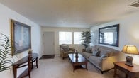 This single-floor space offers a straightforward and functional layout designed for everyday comfort. The living room serves as the heart of the home, featuring wall-to-wall carpeting in a warm neutral tone that adds a sense of coziness underfoot. Smooth, light-colored walls pair with a textured popcorn ceiling to create a classic interior aesthetic. Natural light filters in through a pair of windows along the front wall, brightening the space and offering a view of the surrounding outdoor greenery. A paneled entry door provides convenient direct access to the exterior. The room is well-proportioned, comfortably accommodating a full sofa, an accent armchair, and a dark wood coffee table, which together form a cohesive and inviting seating arrangement. A large decorative mirror mounted on the wall adds depth and visual interest to the space. Subtle details, such as the decorative plant in the corner and coordinating throw pillows, contribute to a lived-in yet tidy atmosphere. The overall ambiance is calm and neutral, making the space highly adaptable to a variety of furnishing styles and personal tastes.