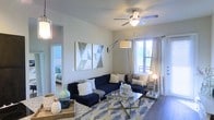This single-floor residence features a thoughtfully designed living area that serves as the centerpiece of the home. The space is finished with smooth, neutral-toned walls that provide a clean backdrop, complemented by warm hardwood-style flooring that adds texture and depth throughout. A ceiling fan with an integrated light fixture anchors the room overhead, contributing both functionality and a polished aesthetic.

Natural light filters in through a large window dressed with sheer and panel curtains, offering a view of an outdoor balcony or patio area beyond. An articulating arc floor lamp and a soft globe lamp add layered ambient lighting, enhancing the room's warm and inviting atmosphere.

The living area is styled with a bold navy sectional sofa, a geometric-patterned area rug in blue and white tones, and a glass-and-gold coffee table, all of which contribute to a contemporary, coordinated aesthetic. Abstract canvas wall art adds a decorative focal point to the main wall.

A glimpse through an open doorway reveals an adjoining bedroom, indicating a well-connected, open floor plan that maximizes the sense of space and flow throughout the residence.