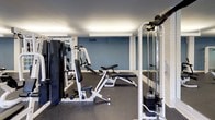 This spacious fitness center is designed to accommodate a full range of workout routines, offering an impressive array of both strength training and cardiovascular equipment. The layout is open and thoughtfully organized, with durable dark rubber flooring throughout that provides comfort and safety during exercise.

The strength training area is well-stocked with multiple cable machines, weight stations, adjustable benches, and resistance equipment, offering users a comprehensive selection for targeting all major muscle groups. Full-length mirrors along one wall enhance the sense of space while allowing users to monitor their form during workouts.

The cardio zone is lined along the windowed wall and features treadmills, elliptical machines, a stationary bike, and a rowing machine. A row of evenly spaced windows fills the space with natural light and provides views of an outdoor pool and courtyard, creating an uplifting atmosphere during workouts.

The facility is illuminated by sleek, linear overhead lighting that ensures bright, even coverage throughout. The walls are finished in a clean combination of soft blue and neutral tones, lending the space a modern, energetic feel. The space is accessed through elegant French doors, adding a refined architectural touch. Overall, this fitness center delivers a well-rounded, functional, and inviting environment for exercise.