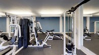 This spacious fitness center is designed to accommodate a full range of workout routines, offering an impressive array of both strength training and cardiovascular equipment. The layout is open and thoughtfully organized, with durable dark rubber flooring throughout that provides comfort and safety during exercise.

The strength training area is well-stocked with multiple cable machines, weight stations, adjustable benches, and resistance equipment, offering users a comprehensive selection for targeting all major muscle groups. Full-length mirrors along one wall enhance the sense of space while allowing users to monitor their form during workouts.

The cardio zone is lined along the windowed wall and features treadmills, elliptical machines, a stationary bike, and a rowing machine. A row of evenly spaced windows fills the space with natural light and provides views of an outdoor pool and courtyard, creating an uplifting atmosphere during workouts.

The facility is illuminated by sleek, linear overhead lighting that ensures bright, even coverage throughout. The walls are finished in a clean combination of soft blue and neutral tones, lending the space a modern, energetic feel. The space is accessed through elegant French doors, adding a refined architectural touch. Overall, this fitness center delivers a well-rounded, functional, and inviting environment for exercise.