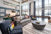 Located at 3196 US-280 E in Birmingham, Alabama, this community clubhouse at Tributary presents a thoughtfully designed gathering space defined by soaring double-height ceilings, floor-to-ceiling windows, and an open, airy floor plan that allows natural light to flood the interior throughout the day.

The entry foyer features a striking geometric-patterned tile floor, a large marble-topped reception desk, warm wood-paneled walls, and an oversized nature-inspired mural, all illuminated by dramatic pendant lighting. A convenient coffee bar with stone countertops and cabinetry is positioned nearby, alongside a staircase with exposed wood beam detailing overhead.

The expansive main lounge area is anchored by a full-height stone fireplace surround with built-in teal shelving and a mounted television. Multiple seating arrangements—including sofas, accent chairs, and casual dining groupings—are spread across wood-look tile flooring and defined by natural fiber area rugs. Decorative candle-style chandeliers add warmth throughout.

A dedicated game area features a shuffleboard table and bistro seating, framed by the same soaring windows that offer views of the surrounding outdoor amenity spaces. Teal shiplap accent walls, exposed wood ceiling beams, and a neutral palette of whites and warm tones unify the space with a refined coastal-contemporary aesthetic.