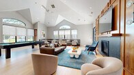 Located at 2101 Glen Dr in Cedar Knolls, NJ, this contemporary amenity lounge is an open-concept gathering space defined by dramatic vaulted ceilings that rise to an impressive peak, accentuated by a large arched window that floods the room with natural light. Light hardwood flooring runs throughout, complementing the soft gray and white color palette that gives the space a clean, modern aesthetic.

The main lounge area centers around a sleek fireplace framed by a striking floor-to-ceiling wood accent wall, which also houses a large flat-screen television. Comfortable seating — including rounded accent chairs and a plush sofa — surrounds a sculptural white coffee table atop a vibrant teal area rug, creating an inviting social hub. A ceiling fan overhead adds both function and style.

A full-size pool table anchors an entertainment zone, while a built-in wet bar and kitchenette with dark countertops and white cabinetry provides convenient service space. A casual dining nook lines one wall with built-in banquette seating and café-style tables, positioned directly beside floor-to-ceiling windows that overlook an outdoor pool area. Glass doors provide direct exterior access, seamlessly connecting indoor and outdoor living. Recessed and track lighting complement the abundant natural light throughout.