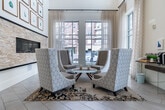 This elegant single-floor lobby and lounge space is defined by its dramatic double-height ceilings, which lend an impressive sense of openness and grandeur. The main lounge area centers around a striking stacked-stone fireplace wall, accented by a grid of framed botanical artwork that extends from floor to ceiling, creating a sophisticated focal point. Light-toned large-format tile flooring runs throughout, reflecting the abundant natural light that pours in through floor-to-ceiling windows dressed with sheer linen curtains. Comfortable high-back upholstered chairs are arranged around a round white coffee table atop a patterned area rug, providing an inviting seating area. A built-in floating shelf along one windowed wall serves as a convenient coffee station, adding a practical and welcoming touch. Adjacent to the main lounge, a glass-walled room enclosed by sleek floor-to-ceiling glazing offers a semi-private workspace or meeting area, featuring a warm wood accent wall and ample table seating. Tall white rectangular planters filled with lush greenery line the corridor, enhancing the fresh, contemporary atmosphere. The overall design blends clean, modern architecture with warm, curated furnishings to create a polished and functional communal environment.