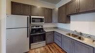 Located at 139 Alexander Bank Dr. in Mooresville, NC, this single-story residence offers a well-designed layout with a clean, neutral aesthetic throughout. The kitchen serves as a functional centerpiece, showcasing dark taupe shaker-style cabinetry, granite-look countertops, a stainless steel double-basin sink, and a full suite of stainless steel appliances including a refrigerator, electric range, over-the-range microwave, and dishwasher. Crown molding adds a refined touch to the kitchen and adjoining living area, which transitions seamlessly between rich hardwood-style flooring and plush carpeting. The living space is bright and open, with recessed lighting complementing the natural light that flows through the windows. The bedrooms are generously proportioned, finished with wall-to-wall carpet and large windows fitted with blinds that frame views of the surrounding greenery. The full bathroom features a tub-shower combination, a dark-stained vanity with a white countertop, hardwood-style flooring, and an adjacent walk-in closet with built-in wire shelving. A door in one of the rooms provides access to an outdoor space, extending the living area beyond the interior. The overall palette of warm beige walls and white trim creates a light, welcoming atmosphere throughout.
