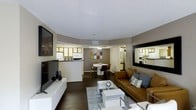 This single-floor residence showcases an open-concept design that seamlessly connects the living, dining, and kitchen areas into one cohesive and functional space. The living area is anchored by a sleek, low-profile media console with a large flat-screen television, complemented by a large wall-mounted mirror that enhances the sense of depth and light. A tan leather sofa adorned with decorative throw pillows provides comfortable seating, centered around a modern white coffee table atop a light area rug. Dark hardwood flooring runs throughout, creating a rich contrast against the warm, neutral-toned walls. The dining area features a round white table with contemporary chairs, lit by a stylish ceiling fixture that adds a refined touch. Just beyond, the kitchen is equipped with light wood cabinetry, white appliances, and a breakfast bar that opens to the main living space — ideal for casual dining or entertaining. The textured ceiling and recessed ventilation details reflect thoughtful construction throughout. Decorative accents, including wall art and sculptural elements, lend personality and warmth to the overall ambiance, making this a practical yet stylish living environment.