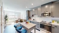 This single-floor residence showcases a thoughtfully designed open-concept layout with modern finishes and a cohesive contemporary aesthetic. The kitchen serves as the heart of the home, featuring sleek gray flat-front cabinetry, a classic white subway tile backsplash, quartz countertops, and a full suite of stainless steel appliances including a gas range, built-in microwave, dishwasher, and French door refrigerator. A central kitchen island with a marble-look quartz surface and seating for two provides both additional prep space and a casual dining option. Wood-look flooring flows throughout, tying the spaces together seamlessly. The living area benefits from large windows that invite abundant natural light and offer views of the surrounding cityscape. A secondary flexible room with generous windows also functions well as a home office or sitting area. Both bedrooms are well-proportioned, each featuring large windows with outdoor views, allowing natural light to fill the rooms. The bathroom is finished with a floating wood-tone vanity, white quartz countertop, black-framed mirror, and wood-look tile flooring. Throughout the residence, recessed lighting, clean white walls, and modern hardware contribute to a bright, polished atmosphere.