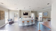 Located at 7030 Watchman Cir in Montgomery, Alabama, this single-floor community clubhouse offers a well-appointed and inviting gathering space designed for both social and functional use. The open layout is anchored by a curved, white quartz-topped island bar, illuminated by pendant lighting and flanked by industrial-style adjustable bar stools — creating an ideal hub for casual interaction. Behind the island, a textured mosaic tile accent wall frames a wall-mounted flat-screen TV above a row of white cabinetry, adding a stylish focal point to the space. The flooring throughout features large-format dark tile laid in a herringbone pattern, providing a sophisticated contrast to the soft gray walls and crisp white crown molding and baseboards. Multiple high-top tables with bar seating are positioned near large white-framed windows, which flood the interior with natural light and offer views of the surrounding exterior. A dedicated area toward the back includes a full-size refrigerator and direct door access to the outdoor pool, making it highly functional for community events. A separate office or workroom is accessible via a doorway fitted with a framed mirror, providing a subtle visual extension of the space. The textured ceilings, recessed lighting, and cohesive neutral palette contribute to a clean, modern, and welcoming ambiance throughout.