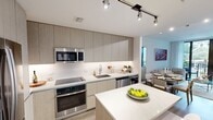 Located in Miramar, FL, this thoughtfully designed unit offers a comfortable and contemporary living experience across a single floor. The open-plan kitchen and living room serve as the heart of the space, maximizing functionality and flow. The kitchen is outfitted with sleek, light wood flat-panel cabinetry extending to the ceiling, stainless steel appliances including a French door refrigerator, built-in oven, microwave, dishwasher, and a wine cooler. A central island with a clean white quartz countertop provides additional workspace and casual seating. Light wood-style flooring runs throughout, lending warmth to the modern aesthetic. The living area is bright and airy, featuring floor-to-ceiling glass doors that open onto a private balcony overlooking a lush, resort-style courtyard complete with a large pool, palm trees, and tropical landscaping. Natural light fills the space generously. The bathroom features a clean, all-white tiled tub-shower combination with a rainfall showerhead, maintaining a crisp, minimal design. A dedicated closet provides practical storage. The overall ambiance is contemporary and polished, ideal for those seeking a well-appointed residence in a vibrant South Florida community.