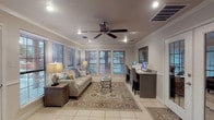 This single-story clubhouse and business center offers a thoughtfully designed communal space that balances comfort with functionality. The main lounge area is bright and welcoming, featuring large windows dressed with horizontal blinds that frame views of the outdoor pool area, allowing generous natural light to filter through. Crown molding lines the ceiling throughout, adding a refined architectural touch, while a ceiling fan with recessed lighting overhead ensures a comfortable atmosphere. Light tile flooring anchors the space, complemented by a traditional area rug beneath a seating arrangement centered around a glass-topped coffee table and a neutral-toned sofa accented with decorative pillows. Adjacent to the lounge, a dedicated business center workstation with a granite countertop and bar-style seating provides computer access for residents or guests. A separate kitchen area is fully equipped with gray raised-panel cabinetry, granite countertops, black appliances including a refrigerator, range, microwave, and additional countertop appliances. French doors lead to an exterior entrance, enhancing accessibility. The overall ambiance is polished yet inviting, making this space well-suited for both productive work sessions and relaxed social gatherings.