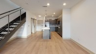 This two-story residence showcases a clean, contemporary design with light wood-style flooring throughout the main living areas and a neutral color palette that enhances the sense of openness. The main floor centers around a well-appointed kitchen equipped with dark wood-grain cabinetry, stainless steel appliances, a tiled backsplash, and a substantial island with a white quartz countertop and pendant lighting overhead. A sleek staircase with metal cable railings connects the two levels, accompanied by an open living area featuring in-wall speakers and a sliding barn door. The upper floor hosts carpeted bedrooms, including one with impressively tall ceilings and multiple windows that allow abundant natural light. Each bedroom includes a dedicated closet with built-in shelving and hanging rods. Two full bathrooms are present, each featuring dark wood-grain vanities with white quartz countertops. The primary bathroom includes a glass-enclosed walk-in shower with tile surround and a separate walk-in closet with open shelving, while the secondary bathroom also connects to a walk-in closet. Recessed lighting is consistent throughout, contributing to the space's bright, modern ambiance.