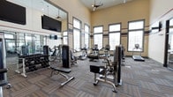 This fitness center offers a spacious, single-level layout designed to accommodate a wide range of workout needs. The room is defined by soaring ceilings that create an open, airy atmosphere, enhanced by large multi-pane windows along the walls that flood the space with natural light. Ceiling fans are installed overhead to maintain comfortable airflow throughout the room.

The facility is outfitted with a diverse selection of strength training machines, including cable and resistance equipment, as well as a dedicated free weights area featuring a dumbbell rack. Cardio machines such as ellipticals and stationary bikes are positioned throughout, providing ample options for aerobic exercise. A flat-screen television is wall-mounted for entertainment during workouts.

Full-length mirrors line one wall, visually expanding the space while allowing users to monitor their form. The flooring consists of durable, patterned carpet tiles suited for a high-traffic fitness environment. Decorative horizontal striping in muted tones adds a structured, finished look to the warm beige walls, contributing to a clean, professional ambiance throughout the facility.