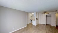 This single-floor space offers a straightforward and functional layout designed for comfortable everyday living. The main living area is open and well-proportioned, featuring light hardwood-style flooring that runs throughout, contributing to a bright and cohesive feel. Neutral beige walls and white baseboards keep the interior palette clean and versatile, while a textured popcorn ceiling adds a classic touch overhead.

A ceiling fan is positioned in the living area, providing both airflow and ambient lighting. The kitchen is partially open to the living space, separated by a half-wall counter that serves as a natural dividing element while maintaining visual connectivity. Inside the kitchen, white cabinetry lines the walls and is paired with white appliances, including a range and washer, creating a tidy, uniform appearance. An overhead fluorescent light fixture illuminates the workspace effectively.

A doorway from the living area leads to additional spaces within the unit, and a closet door is visible near the kitchen area, offering convenient storage. Overall, the space presents a practical and well-maintained interior with a neutral, move-in-ready aesthetic suitable for a range of occupants.