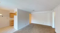This single-floor residence offers a functional and straightforward layout designed for comfortable everyday living. The main living area is generously sized, featuring neutral-toned wall-to-wall carpeting and crisp white walls that create a clean, bright atmosphere. A sliding glass door floods the space with natural light and provides direct access to the outdoors, while a half-wall partition visually separates the living area from the adjoining kitchen zone.

The galley-style kitchen is well-appointed with warm honey-toned wood cabinetry lining both walls, offering ample upper and lower storage. Light-colored countertops provide practical workspace, and the kitchen is equipped with a range, refrigerator, dishwasher, and a double stainless steel sink. Light tile flooring runs throughout the kitchen, complemented by a pendant chandelier that adds a touch of warmth to the dining area just beyond.

The bedrooms are carpeted and bathed in natural light from large windows. One bedroom features a spacious reach-in closet with a full-width hanging rod, providing excellent storage capacity. The bathroom is fully tiled from floor to ceiling in white, with a clean vanity, mirrored medicine cabinet, and a bright, utilitarian design. Throughout the space, the consistent use of neutral tones and practical finishes creates a cohesive, move-in-ready environment.