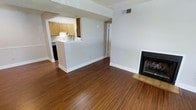 This single-floor residence offers a practical and comfortable layout designed to maximize the sense of openness throughout. The main living area is anchored by rich, warm-toned hardwood flooring that extends throughout the space, creating a cohesive and inviting atmosphere. A classic fireplace with a clean surround serves as a focal point in the living room, adding a cozy element to the space.

The kitchen is thoughtfully positioned adjacent to the living area and connected via a pass-through opening with a raised breakfast bar, making it ideal for casual dining and easy interaction between spaces. The kitchen features warm wood cabinetry, a sink positioned beneath the pass-through window, and space for appliances including a range. The white walls and neutral finishes throughout the space keep it feeling bright and airy.

A hallway visible from the main living area leads to additional rooms, providing a clear and functional flow through the home. Textured ceilings and visible HVAC vents speak to the practical infrastructure of the residence. The combination of the hardwood floors, fireplace, and open kitchen layout gives this space a welcoming character that balances functionality with everyday comfort.