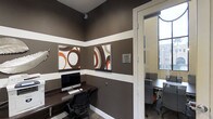 Located at 1935 Northstar Way in San Marcos, this business center offers a thoughtfully designed single-floor workspace that blends functionality with contemporary style. The interior features a rich, earthy color palette with deep taupe walls accented by crisp white chair-rail striping, creating a polished and professional atmosphere. Bold, graphic artwork in coordinating brown, rust, and cream tones adds a modern touch to the walls, reinforcing the space's design-forward aesthetic. A dedicated computer workstation is equipped with an iMac and ergonomic seating, providing a well-appointed setup for individual work or research tasks. An adjacent windowed area, partially visible through a glass-paneled door, offers additional seating and workspace with natural light filtering in from exterior windows — enhancing the overall brightness and openness of the environment. The use of dark wood furnishings and task-oriented chairs throughout contributes to a cohesive, professional look. Whether used for remote work, meetings, or general business tasks, this business center is a well-equipped, stylishly appointed space suited to a variety of professional needs.