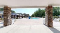 Located at 1100 Cannonball Run in Knightdale, NC, this single-level outdoor amenity space is designed for resort-style community living. At its centerpiece is a generously sized, freeform swimming pool with a shallow sun shelf and ample surrounding deck space lined with lounge chairs, perfect for relaxing under the North Carolina sun. A covered pavilion anchors the space, featuring distinctive stone columns and a beadboard ceiling. Inside the pavilion, a spacious granite-topped bar island with seating for multiple guests serves as a social hub, complemented by pendant lighting, a ceiling fan, wall-mounted television, and direct sightlines to the pool. An adjacent covered lounge area offers plush sectional seating with vibrant orange and navy cushions beneath an elegant chandelier, creating a stylish gathering space. A stone outdoor fireplace with built-in bench seating and a mounted television adds warmth and character to the covered terrace. Beyond the main structure, a dedicated fire pit area with Adirondack chairs is nestled against a lush, wooded backdrop, providing an intimate outdoor retreat. Together, these features create a comprehensive and well-appointed outdoor entertaining environment.