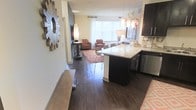 This single-floor residence showcases an open-concept layout that connects the kitchen and living areas in a seamless, functional design. Rich, dark hardwood flooring runs throughout, providing a warm and cohesive foundation that ties the spaces together.

The kitchen is a standout feature, equipped with dark espresso cabinetry, light granite countertops, and stainless steel appliances. A peninsula with bar seating serves as both a prep space and a casual dining area, illuminated by a decorative pendant chandelier that adds an elegant focal point overhead. A subway tile backsplash completes the kitchen's polished aesthetic.

The living area benefits from generous natural light streaming through large windows dressed with horizontal blinds. The space is further enhanced by a decorative sunburst mirror mounted on the wall near the entryway, adding a stylish artistic element. Neutral wall tones complement the warm flooring and furnishings throughout.

Overall, the space strikes a balance between modern design elements and comfortable livability, with an open flow that maximizes the sense of space and encourages easy movement between functional areas.