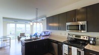 This single-floor space showcases a thoughtfully designed open-concept layout that seamlessly connects the kitchen and living area. The kitchen is a standout feature, outfitted with sleek dark cabinetry that contrasts elegantly against light countertops. Stainless steel appliances — including a built-in microwave, electric range, and dishwasher — lend a contemporary, polished look. An L-shaped counter configuration maximizes workspace and flow, while a double sink with pendant lighting overhead adds both functionality and style. The pendant light fixture, featuring multiple frosted glass shades, serves as a refined focal point above the kitchen peninsula. Beyond the kitchen, the living area benefits from generous natural light streaming through large windows, creating a bright and airy atmosphere. The open sightlines between the kitchen and living space enhance the sense of roominess throughout. Clean white walls and consistent flooring tie the spaces together cohesively. Overall, the unit presents a modern, functional aesthetic well-suited for comfortable everyday living.