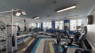 Located at 7029 W Friendly Ave in Greensboro, North Carolina, this well-appointed fitness center offers a comprehensive exercise facility designed to support a wide range of workout routines. The space is spread across a single floor and is organized into distinct functional zones, each catering to different training needs.

The main cardio area is generously sized and lined with an impressive array of treadmills, elliptical machines, stair climbers, and stationary bikes. Large windows along the perimeter flood the space with natural light, while bold blue accent walls and vibrant multi-toned striped carpet tiles add a dynamic, energizing aesthetic throughout. Recessed and overhead panel lighting ensures bright, even illumination. Full-length mirrors along one wall enhance the sense of space and allow users to monitor their form.

A dedicated strength training zone features a multi-station cable machine, a weight bench with rubber floor matting, and a wall-mounted television for added convenience. An adjoining studio space is equipped with indoor cycling bikes, a mirrored wall, a barre-style rail, and an organized rack of kettlebells and free weights in varying sizes. Stability balls, BOSU trainers, and resistance accessories are neatly stored along the studio walls.

The cohesive design, ample equipment variety, and abundance of natural light make this fitness center a practical and motivating environment for users of all fitness levels.