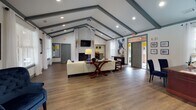 Located at 377 Athens Way in Nashville, Tennessee, this single-story community or leasing center offers a well-appointed and welcoming interior designed for both comfort and functionality. The space is defined by its striking vaulted ceilings accented with bold gray decorative beams, which lend an architectural character that carries throughout the entire floor plan. Recessed lighting and ceiling fans complement the natural light that streams in through multiple white-framed windows dressed with sheer curtains and wood blinds.

The main lounge area serves as the heart of the space, centered around a white-painted brick fireplace with a mounted flat-screen television above. A cream sectional sofa, vibrant green area rug, and a wooden coffee table create an inviting gathering spot. Adjacent to the lounge, a seating nook features a pair of tufted navy blue armchairs arranged around a side table, offering a more intimate setting with views of the outdoor surroundings.

A dedicated private office area includes a substantial executive desk and leather chair beneath the same vaulted ceiling, with interior windows providing a visual connection to the lounge. Rich hardwood-style flooring flows seamlessly throughout, unifying the open layout. Curated artwork and decorative accents add warmth and a polished, professional atmosphere to this thoughtfully designed space.