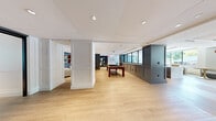 This thoughtfully designed single-floor amenity space offers a generous and versatile layout across multiple distinct functional areas, all unified by a refined contemporary aesthetic. Light hardwood flooring flows throughout, complemented by white paneled walls, recessed lighting, and a neutral palette punctuated by slate-blue cabinetry and warm brass accents.

The centerpiece of the space is a striking kitchen and bar area, featuring an expansive quartz-topped island, herringbone hardwood flooring underfoot, a warm wood-paneled ceiling, and oversized brass pendant lights that lend an air of sophistication. Adjacent, a spacious lounge area invites relaxation with plush leather and upholstered seating arranged around a marble fireplace flanked by dark paneled built-ins with gold-lit display niches and a wall-mounted screen.

A dedicated game room accommodates a billiards table and is defined by its open, airy layout with views toward glass-partitioned areas beyond. The space also includes intimate meeting and collaboration rooms, each appointed with white tables, curated artwork, and statement pendant lighting. A smaller breakout nook features built-in shelving with warm amber interiors and gold tulip tables. Large windows throughout flood the space with natural light and offer lush, tree-lined outdoor views, creating a bright and welcoming atmosphere throughout.