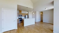 This single-floor residence showcases a thoughtfully designed open-concept layout that seamlessly connects the kitchen and living areas, creating a spacious and airy atmosphere. Soaring vaulted ceilings amplify the sense of openness, while warm hardwood-style flooring flows continuously throughout the main living space, lending a cohesive and inviting feel.

The kitchen is a standout feature, equipped with stainless steel appliances including a gas range, microwave, and refrigerator. Warm-toned flat-front cabinetry provides ample storage, and a central island with a clean white finish serves as both a prep space and a casual gathering point. Elegant pendant lighting hangs above the island, complementing the track lighting positioned over the main cooking area.

The open living area beyond the kitchen benefits from the elevated ceiling height, offering a generous and flexible space. Neutral light-toned walls and abundant recessed and ceiling-mounted fixtures ensure the interior remains bright and welcoming. A hallway visible from the main living area leads to additional rooms, with interior doors finished in crisp white. The overall design strikes a balance between modern functionality and comfortable livability, making it a well-rounded and practical living space.