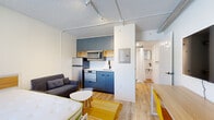 Located at 1104 Avondale Circle in Nashville, TN, this compact yet efficiently designed unit offers a thoughtful single-floor layout that maximizes every inch of available space. The open-plan main area seamlessly combines sleeping, living, and dining functions, finished with warm light wood flooring and crisp white walls that keep the space feeling bright and airy. A window with blinds allows natural light to filter in, enhancing the overall sense of openness. Exposed track lighting and visible ductwork along the textured ceiling lend the space an industrial-modern character. The kitchenette is a standout feature, equipped with sleek blue-gray cabinetry, open wood shelving, a stainless steel refrigerator, microwave, range, and a single-basin sink set against a herringbone tile backsplash. A wall-mounted TV and a dedicated desk with seating offer both entertainment and workspace functionality. The private bathroom is clean and efficiently arranged, featuring a walk-in shower with curtain, a wall-mounted sink, a framed mirror with black hardware accents, and light wood flooring with a slatted wood bath mat — delivering a minimal yet modern aesthetic throughout.