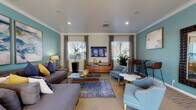 Located at 1400 Stone Pine Terrace in Fremont, California, this single-floor communal lounge space offers a well-appointed and thoughtfully designed interior. The space is defined by its signature teal accent walls, which are complemented by neutral beige tones and crisp crown molding throughout, creating a cohesive and contemporary aesthetic.

The main lounge area is generously sized, featuring a large sectional sofa in charcoal gray, accented with navy and yellow throw pillows that add a pop of color. A wall-mounted flat-screen television sits atop a warm wood media console, flanked by multiple windows dressed in gray drape panels that allow abundant natural light to filter in. Recessed lighting overhead ensures the space is well-lit at all times.

A variety of seating arrangements encourages both relaxation and social interaction, including mid-century-inspired accent chairs in soft blue and gray, marble-top side tables, and a bistro-style high-top table with leather counter chairs. Decorative wall art, a sculptural mirror, and lush indoor plants add character and warmth throughout.

A separate nook features a solid wood standing work table with leather-upholstered chairs, ideal for casual meetings or focused tasks. Glass entry doors connect the interior to the outdoors, enhancing the sense of openness and accessibility.