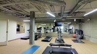 This fitness center offers a comprehensive workout environment designed to accommodate a variety of exercise needs. The space features an industrial aesthetic, with exposed ductwork, concrete support columns, and open ceiling infrastructure that lend it a raw, utilitarian character. The flooring consists of light hardwood, providing a clean and durable surface throughout the workout area, complemented by rubber mats in designated exercise zones.

The cardio section is well-appointed with multiple treadmills, an elliptical trainer, and a stationary bike, catering to a range of cardiovascular workout preferences. A wall-mounted flat-screen television is positioned for viewing during workouts. Strength and functional training areas include a weight bench, free weights, and a stability ball, offering options for resistance and flexibility training.

Full-length mirrors line one wall, visually expanding the space and allowing users to monitor their form during exercise. Fluorescent lighting ensures the space is well-lit throughout. The layout is thoughtfully organized to provide sufficient room between equipment, promoting safe and comfortable use. Overall, this fitness center combines practical functionality with an industrial-modern aesthetic.