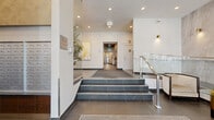 The Wesley Place lobby at 1041 Comox St in Vancouver's West End offers a refined and welcoming first impression. The space is thoughtfully designed across two interconnected hallway areas, combining contemporary architecture with upscale finishes throughout. Large-format grey porcelain floor tiles flow seamlessly from the main entrance through the hallways, complemented by a branded "Wesley Place" entrance mat that greets residents and guests upon arrival. Floor-to-ceiling glazing at the front of the lobby allows abundant natural light to pour in, offering views of the tree-lined street outside. A dramatic floor-to-ceiling marble-clad accent wall anchors the seating lounge, where a curved charcoal sofa, glass-top side tables with gold accents, and a lush indoor plant create an inviting atmosphere. A statement chandelier with gold arms and candelabra-style lighting adds an elegant focal point overhead. A glass-and-steel staircase with illuminated steps connects the entry level to the inner hallway, which features a decorative gold botanical artwork on white walls. Recessed and wall-mounted gold sconce lighting throughout enhances the warm, sophisticated ambiance. Resident mailboxes and elevator access with wood-panel detailing are conveniently located within the inner hallway, completing this polished and functional lobby space.