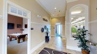 This single-floor amenity space is designed around a central hallway that serves as the main artery connecting multiple functional areas. The corridor features light-toned ceramic tile flooring accented by a decorative area rug, while warm beige walls are finished with white chair-rail molding and crown molding that extend toward notably elevated ceilings. Recessed lighting illuminates the space evenly throughout.

To the left, a community room is accessible through a wide doorway, identifiable by its signage and furnished with a pool or billiards table, offering a dedicated space for recreation and social gatherings. To the right, a fitness area is visible through an arched window opening, equipped with exercise equipment including a stability ball and cardio machines, bathed in bright overhead lighting.

Decorative wall art, including hexagonal pieces and a floral arrangement atop a small dark console table, adds visual interest along the hallway. At the far end, a glass-paneled door allows natural light to filter in, enhancing the open and airy atmosphere. The overall design balances functionality with an approachable, well-maintained aesthetic suitable for a shared amenity facility.