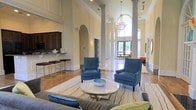 This sophisticated space makes an immediate impression with its dramatic double-height ceilings and stately round columns that anchor the main living area. A crystal chandelier hangs as a centerpiece, drawing the eye upward toward the grand arched windows that flood the interior with abundant natural light. The flooring transitions seamlessly between rich hardwood and polished tile, adding texture and warmth throughout.

The main lounge area is thoughtfully appointed with plush blue accent chairs arranged around a marble-topped coffee table, all set atop a patterned area rug. Crown molding, wainscoting, and decorative arched doorways reinforce the classical architectural character of the space. A built-in wet bar or kitchenette area sits to one side, complete with dark cabinetry, granite countertops, and bar-height seating with sleek gold-accented stools — ideal for entertaining or casual gatherings.

Toward the rear, large glass entry doors open to an outdoor area, offering a seamless connection to the exterior and enhancing the sense of openness. A decorative round mirror and carefully placed lighting fixtures contribute to the polished, upscale ambiance. Overall, this space balances classical elegance with inviting comfort, making it well-suited for social gatherings and community interaction.