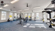 This well-appointed fitness center offers a generous, open workout environment designed to accommodate a full range of exercise needs. The interior is finished with crisp white walls and patterned blue-grey carpet flooring, creating a clean, modern aesthetic throughout. Multiple black ceiling fans are suspended from the white ceiling alongside rows of flush linear light fixtures, ensuring consistent air circulation and illumination across the entire space.

Large white-framed windows line the perimeter walls, flooding the room with natural light and providing exterior views of the surrounding area. The abundance of windows contributes to an airy, open atmosphere that makes the space feel expansive and welcoming.

The facility is thoroughly equipped with a wide selection of cardio machines, including treadmills, ellipticals, stair climbers, and stationary bikes. Strength training equipment includes cable and multi-use weight machines, a dedicated dumbbell rack with a full range of free weights, a functional training rig with suspension straps, and a medicine ball storage rack. Full-length mirrors are mounted along one wall, enhancing the sense of space and supporting proper form during workouts. Potted plants add subtle organic warmth to the otherwise athletic environment.