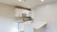Located at 159 W 200 N in Salt Lake City, Utah, this efficiently designed single-floor unit offers a clean, modern aesthetic throughout. The open-concept kitchen serves as the centerpiece of the space, outfitted with crisp white shaker cabinets accented by brushed gold hardware, white quartz countertops, and a full suite of white appliances including a range, microwave, dishwasher, and refrigerator. A peninsula island provides additional prep and dining space, enhancing the functional layout. Light hardwood flooring runs throughout, complementing the neutral warm-gray wall tones and recessed lighting that keep the interior bright and inviting. The living area benefits from excellent natural light, with sliding glass doors opening directly onto a private balcony overlooking a landscaped outdoor area with mature trees — a notable feature that brings the outdoors in. The bathroom is thoughtfully finished with a sliding glass-door walk-in shower, clean white tile surround, and wood-look flooring. Two hallways connect the key areas of the unit seamlessly, while a dedicated closet provides practical storage. The overall design is cohesive and contemporary, emphasizing light, clean lines, and efficient use of space throughout.