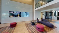This contemporary communal space showcases a sophisticated architectural design characterized by soaring double-height ceilings, expansive floor-to-ceiling glass curtain walls, and large-format wood-toned tile flooring that flows seamlessly throughout. The main lounge area centers around a bold, oversized feature wall clad in glossy white panels housing a large integrated display screen. Seating in this zone includes a curved charcoal sofa and vibrant magenta ottomans arranged over a striking marbled area rug, creating a dynamic visual focal point. A glass-railed mezzanine overlooks this lounge, adding vertical drama to the space. Adjacent to the lounge, a dedicated coworking zone offers long bar-height tables with leather and metal seating, positioned directly in front of floor-to-ceiling windows that frame views of a palm tree-lined outdoor area, flooding the room with natural light. Private office or conference spaces enclosed by glass partitions provide semi-transparent workspaces for focused tasks. The lobby entry features elevator access framed by dark tile accent walls, a white platform seating bench, and a sculptural pendant light installation suspended against a dramatic curtain wall overlooking surrounding streetscapes. The overall ambiance is polished, modern, and professionally appointed.