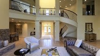 This impressive home makes a striking first impression with its dramatic double-height entry and living area, where soaring ceilings create an immediate sense of grandeur. A sweeping curved staircase serves as the architectural centerpiece, accented by ornate wrought iron balusters that extend along the upper-level balcony overlooking the main floor below. Decorative columns frame the entryway corridor, which leads to a set of elegant French doors that flood the space with natural light and offer a warm welcome from the exterior. The main living area is generously proportioned, featuring large-format tile flooring laid in a classic pattern that grounds the open layout. The upper balcony, wrapped in matching wrought iron railings with detailed geometric motifs, provides an elevated vantage point and adds to the home's sense of layered vertical space. Recessed lighting throughout the ceiling supplements the natural light, ensuring the interiors remain bright and inviting. The living space is styled with a mix of tufted seating and a glass-topped coffee table, reflecting a refined aesthetic. Warm neutral tones on the walls and architectural trim tie the entire space together, conveying an atmosphere of sophisticated comfort and timeless elegance.