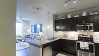This thoughtfully designed single-floor residence showcases a seamless open-concept layout that effortlessly connects the kitchen and living areas, creating a bright and cohesive living environment. The kitchen is a standout feature, boasting rich dark cabinetry that provides striking contrast against light granite countertops. A peninsula with an integrated sink and under-cabinet lighting serves as both a functional workspace and a casual gathering spot. Elegant pendant lights hang above the peninsula, complementing the track lighting overhead for a layered, well-lit atmosphere. Stainless steel appliances, including a dishwasher, complete the kitchen's modern aesthetic. The open living area flows naturally from the kitchen, featuring wood-style flooring that adds warmth and continuity throughout the space. A ceiling fan with light fixture enhances comfort and airflow in the living room, which is filled with natural light filtering in through large windows dressed with sheer curtains. The overall ambiance is contemporary and inviting, with a clean, neutral color palette accented by bold decorative touches that lend personality and charm to every corner of the home.