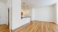 This single-floor unit offers a thoughtfully arranged open-concept layout that combines the living, dining, and kitchen areas into one cohesive space. Warm-toned wood-look flooring extends continuously throughout, lending a sense of visual continuity and warmth across every room.

The living and dining area benefits from elevated ceilings and multiple windows that invite natural light, creating an airy, open feel despite the compact footprint. Track lighting further illuminates the space with a modern touch.

The kitchen is efficiently designed in a galley style and opens to the main living area via a peninsula counter with a dark stone-style surface and bar seating, accented by pendant lighting overhead. Warm wood cabinetry lines the walls, complemented by stainless steel appliances including a refrigerator, electric range, and dishwasher. A deep sink with a modern faucet completes the functional workspace.

The bathroom is clean and straightforward, featuring wood vanity cabinetry, a light countertop, a mirrored medicine cabinet, and Hollywood-style vanity lighting above. Wood-look flooring continues into this space, maintaining design consistency throughout the unit.

Overall, the space presents a bright, neutral aesthetic with functional finishes suited to everyday living.