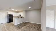 Located at 3325 Oak Forest Dr in Fayetteville, NC, this single-floor unit offers a clean, modern aesthetic defined by neutral warm-gray walls, crown molding, and durable wood-look plank flooring throughout the main living areas. The open-concept layout connects the living and dining areas seamlessly, with the kitchen featuring a peninsula counter with bar seating, ample white shaker-style cabinetry, light stone countertops, and a full suite of black appliances including a refrigerator, gas range, over-the-range microwave, and dishwasher. Natural light filters into the living spaces through windows fitted with horizontal blinds. The bedrooms are finished with plush carpeting, providing a comfortable and quiet retreat, and include generous closet space. Doors throughout feature a classic six-panel design with bright white finishes that contrast cleanly against the neutral walls. The bathroom is well-appointed with wood-look tile flooring, a full soaking tub with shower combination, and clean white fixtures. Recessed and flush-mount lighting illuminates the interior efficiently. The floor plan, as seen in the dollhouse view, offers a functional and well-organized layout with clearly defined living, dining, kitchen, and bedroom zones.