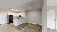 Located at 3325 Oak Forest Dr in Fayetteville, NC, this single-floor unit offers a clean, modern aesthetic defined by neutral warm-gray walls, crown molding, and durable wood-look plank flooring throughout the main living areas. The open-concept layout connects the living and dining areas seamlessly, with the kitchen featuring a peninsula counter with bar seating, ample white shaker-style cabinetry, light stone countertops, and a full suite of black appliances including a refrigerator, gas range, over-the-range microwave, and dishwasher. Natural light filters into the living spaces through windows fitted with horizontal blinds. The bedrooms are finished with plush carpeting, providing a comfortable and quiet retreat, and include generous closet space. Doors throughout feature a classic six-panel design with bright white finishes that contrast cleanly against the neutral walls. The bathroom is well-appointed with wood-look tile flooring, a full soaking tub with shower combination, and clean white fixtures. Recessed and flush-mount lighting illuminates the interior efficiently. The floor plan, as seen in the dollhouse view, offers a functional and well-organized layout with clearly defined living, dining, kitchen, and bedroom zones.