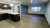 This single-floor space showcases a thoughtfully designed open-concept layout that seamlessly connects the kitchen and living areas. The kitchen features a dark-cabineted island topped with a light quartz or stone countertop, complemented by pendant lighting overhead that adds a modern touch. Warm, wood-style luxury vinyl or laminate flooring runs throughout, creating a cohesive and inviting aesthetic.

The living area is defined by a striking deep blue accent wall that adds a bold contrast to the neutral cream-toned walls and white trim. A large window along the exterior wall fills the space with natural light while offering views of the surrounding neighborhood. A baseboard heating unit runs beneath the window, providing practical climate control.

A hallway visible from the main area leads to additional rooms, with a mirror-paneled door adding depth and light to the corridor. Recessed lighting punctuates the ceiling throughout, ensuring well-distributed illumination. The overall ambiance is clean, modern, and move-in ready, with a neutral palette that provides a versatile backdrop for a variety of interior styles. The open floor plan enhances the sense of spaciousness, making this single-level space feel both functional and welcoming.