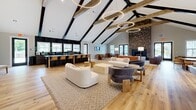 Located at 5226 Whispering Willow Cir in Murfreesboro, TN, this impressive single-floor space offers a generously sized, open-concept layout designed for both comfort and entertainment. The standout architectural feature is the dramatic vaulted ceiling adorned with exposed wood and black-painted beams, which rise to great heights and lend the interior a rustic-modern character. Suspended circular pendant lights add a contemporary touch overhead.

The living area is anchored by a striking floor-to-ceiling brick fireplace that serves as a bold focal point, complemented by a seating arrangement of plush sofas and textured accent chairs arranged atop patterned area rugs. Light hardwood flooring runs throughout, enhancing the warm, inviting ambiance.

A full bar area with dark cabinetry, bar seating, and beverage refrigerators occupies one side of the space, making it well-suited for hosting gatherings. Multiple dining tables with modern chairs are positioned throughout, offering flexible seating arrangements. A pool table adds a recreational element to the open floor plan.

Natural light floods the interior through numerous large windows and glass double doors that open to an outdoor patio, where additional seating is visible. A quieter lounge area with softer furnishings provides a more intimate setting. The overall design blends rustic warmth with modern finishes throughout.