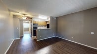 Located at 8550 E Speedway Blvd in Tucson, Arizona, this single-story residence offers a practical and well-organized layout across its living, kitchen, bedroom, and bathroom spaces. The open living area is finished with rich, dark hardwood-style flooring that extends throughout the main living zone, complemented by neutral gray walls and a textured ceiling. A ceiling fan provides additional comfort, while a window at the front of the living area allows natural light to brighten the space. The kitchen is defined by a half-wall peninsula that separates it from the living area, offering a convenient pass-through design. Inside, the kitchen features warm wood-toned cabinetry, a full suite of black appliances including a range, dishwasher, and refrigerator, a double-basin sink, and light-toned countertops. Light vinyl flooring and under-cabinet lighting contribute to a functional workspace. The bedrooms are carpeted for comfort, with neutral tones and textured walls creating a calm atmosphere. The primary bedroom includes a spacious sliding-door closet with dual sections and direct access to the bathroom. The bathroom presents a wood-toned vanity with a granite-style countertop, an inset sink, a large mirror, and brushed nickel fixtures, with additional storage and a shower area nearby. The overall layout is efficient and cohesive throughout.