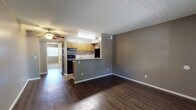 Located at 8550 E Speedway Blvd in Tucson, Arizona, this single-story residence offers a practical and well-organized layout across its living, kitchen, bedroom, and bathroom spaces. The open living area is finished with rich, dark hardwood-style flooring that extends throughout the main living zone, complemented by neutral gray walls and a textured ceiling. A ceiling fan provides additional comfort, while a window at the front of the living area allows natural light to brighten the space. The kitchen is defined by a half-wall peninsula that separates it from the living area, offering a convenient pass-through design. Inside, the kitchen features warm wood-toned cabinetry, a full suite of black appliances including a range, dishwasher, and refrigerator, a double-basin sink, and light-toned countertops. Light vinyl flooring and under-cabinet lighting contribute to a functional workspace. The bedrooms are carpeted for comfort, with neutral tones and textured walls creating a calm atmosphere. The primary bedroom includes a spacious sliding-door closet with dual sections and direct access to the bathroom. The bathroom presents a wood-toned vanity with a granite-style countertop, an inset sink, a large mirror, and brushed nickel fixtures, with additional storage and a shower area nearby. The overall layout is efficient and cohesive throughout.