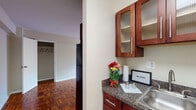This single-floor unit offers a well-organized layout with classic design elements throughout. The kitchen is a standout feature, equipped with rich, dark cherry wood cabinetry accented by frosted glass upper doors and sleek bar-pull hardware. Dark granite-style countertops provide ample prep space, complemented by a stainless steel sink and a gas range. Light ceramic tile flooring in the kitchen and entryway offers a clean, practical surface near the front door.

The main living area is characterized by warm, eye-catching parquet hardwood flooring arranged in a classic geometric pattern, lending the space a timeless and elegant feel. The room is open and versatile, with neutral-toned walls that maximize light. A large window fitted with horizontal blinds and a wall-mounted air conditioning unit ensures year-round comfort, while a radiator provides supplemental heating. A walk-in closet offers generous storage.

The bathroom is finished with white subway tile wainscoting, a pedestal sink with brushed nickel fixtures, a framed mirror, and a shower-tub combination. The overall ambiance of the space is practical and comfortable, with classic architectural details providing lasting character throughout.