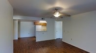 This single-floor residence offers a practical and functional layout designed for comfortable everyday living. The main living area is open and airy, featuring rich, dark hardwood-style flooring that extends throughout the space, adding warmth and a sense of continuity. A ceiling fan with an integrated light fixture is centrally mounted, providing both air circulation and ambient lighting to the room.

The kitchen is positioned adjacent to the living area and is accessible via a pass-through counter, creating a semi-open connection between the two spaces. The kitchen features warm wood-toned cabinetry, a white refrigerator, and an under-cabinet light that brightens the work area. The pass-through design allows for easy interaction between the kitchen and living space while maintaining a degree of separation.

The interior walls are finished in a clean, neutral white, contributing to a bright and simple aesthetic throughout. A standard interior door visible from the living area leads to additional rooms within the unit. Ceiling vents suggest a central HVAC system for year-round climate control. Overall, the space presents a straightforward, well-maintained layout that prioritizes functionality and ease of use.